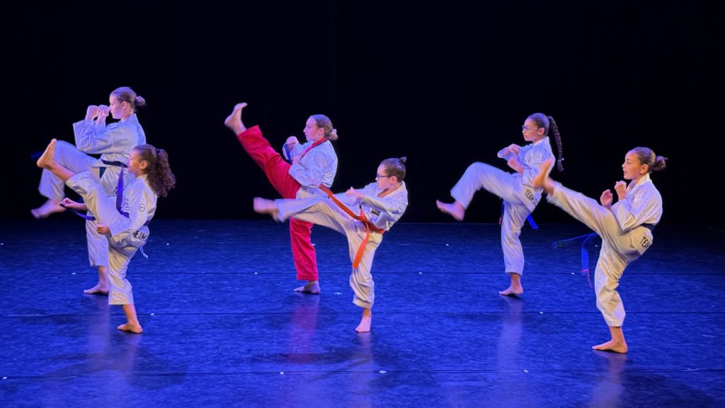 Group of young martial artists in white gis performing high kicks on a dark stage with blue lighting.