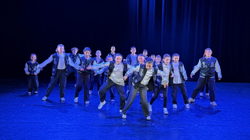 Group of young dancers in matching jackets performing a choreographed routine on a blue-lit stage.