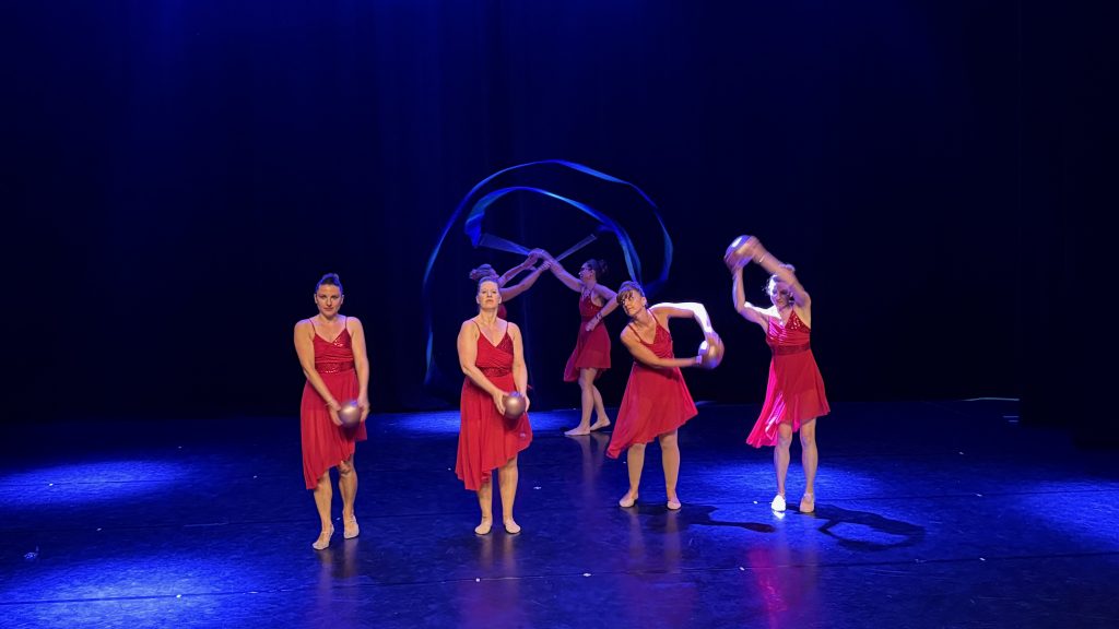 Six dancers in red dresses perform a routine on a dark blue stage, some holding silver balls.