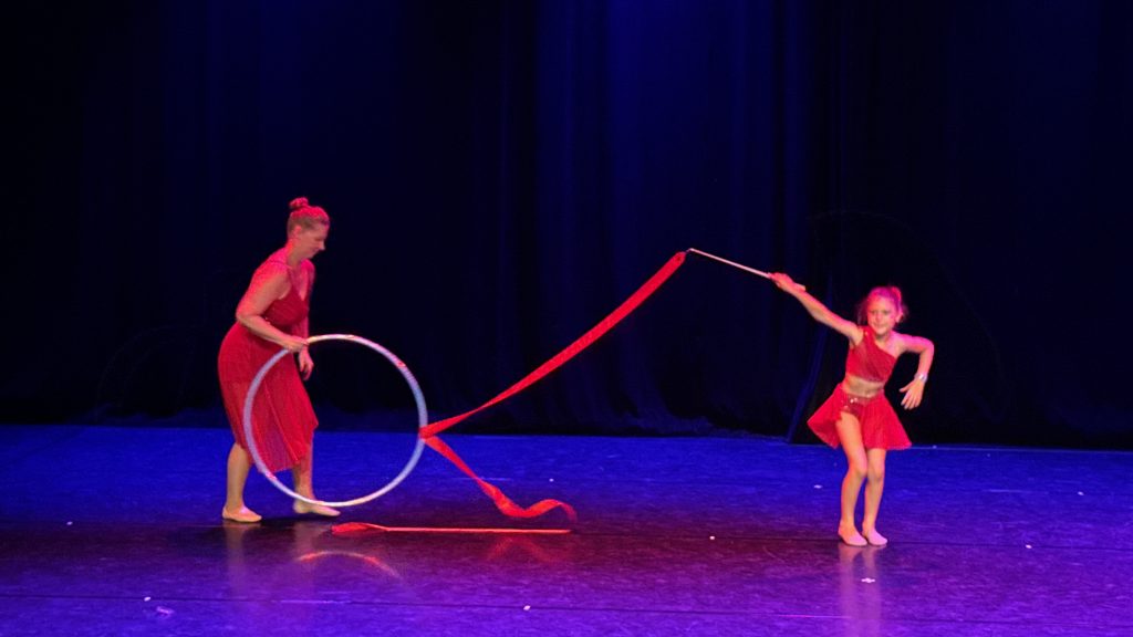 Two performers in red dresses dance on a blue-lit stage; one spins a hoop while the other holds a long red ribbon aloft.