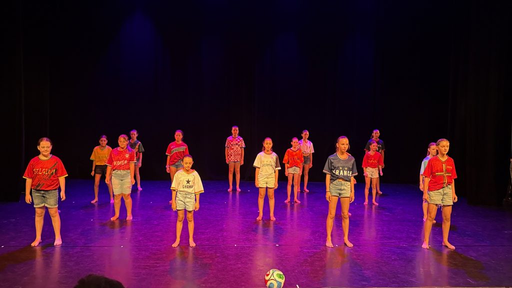 Group of children in colorful jerseys standing on a lit stage with a soccer ball in front, purple backdrop overhead.
