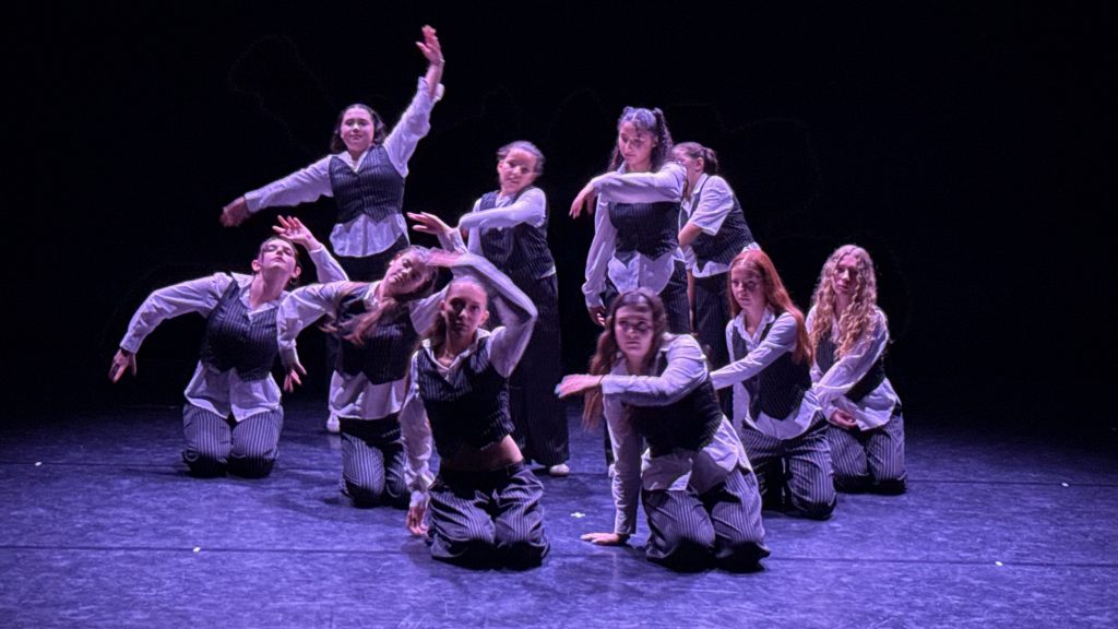 Group of dancers in white shirts and pinstriped vests perform on a dark stage, mid-routine, with dramatic poses in unison.
