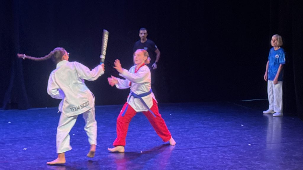 Two young martial artists spar on a blue-lit stage, one in a white gi with a blue belt and the other in a white top with red pants, as a man watches from the back and a girl in a blue shirt stands on the right.