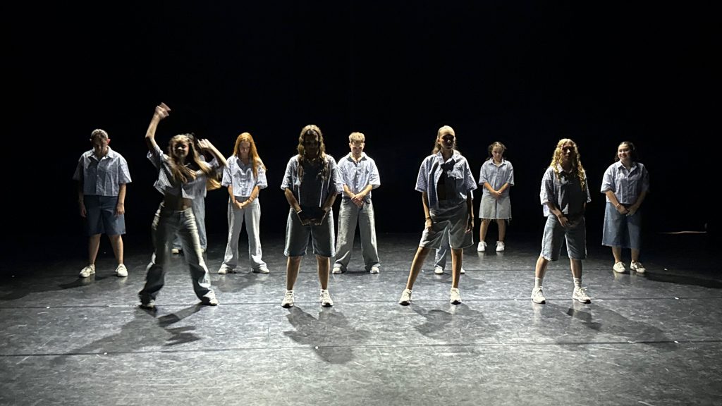 A line of young dancers stands on a dark stage in matching light plaid tops and gray shorts, preparing for a performance.