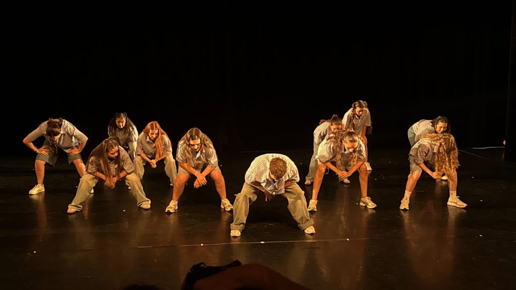 Group of young dancers in checkered shirts kneeling and bending forward on a dark stage.