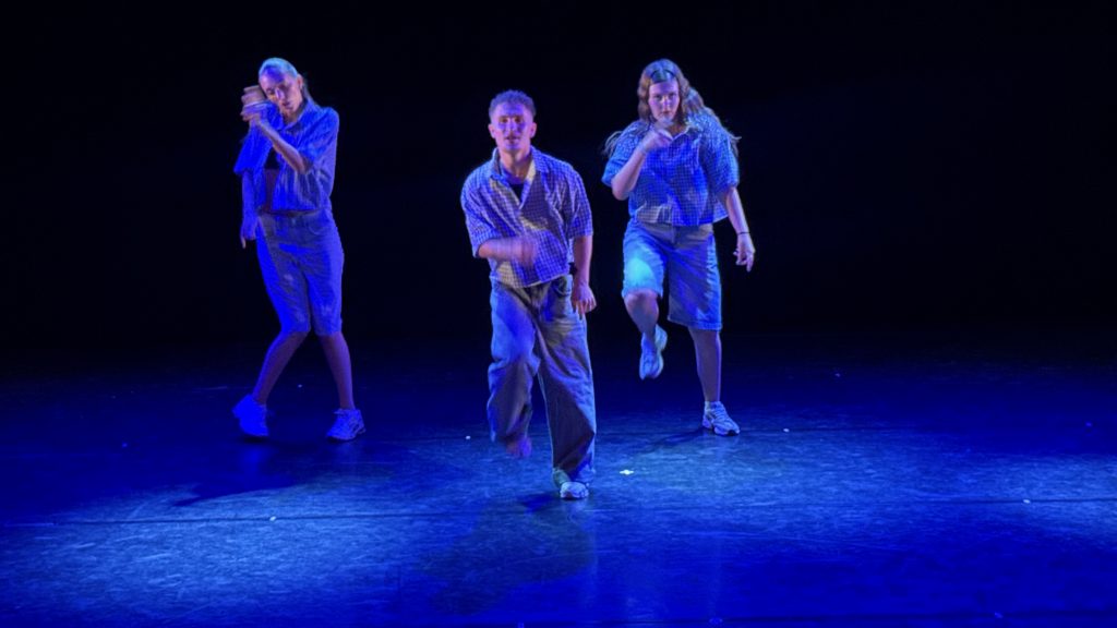 Three dancers in blue plaid outfits perform on a dark stage bathed in blue light, mid-performance. This trio moves in a line, with the center dancer stepping forward.