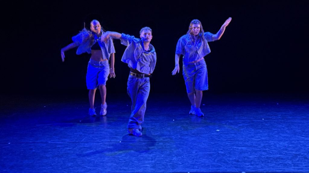 Three dancers perform a blue-lit routine on a dark stage, wearing casual denim and plaid shirts with arms raised in motion.